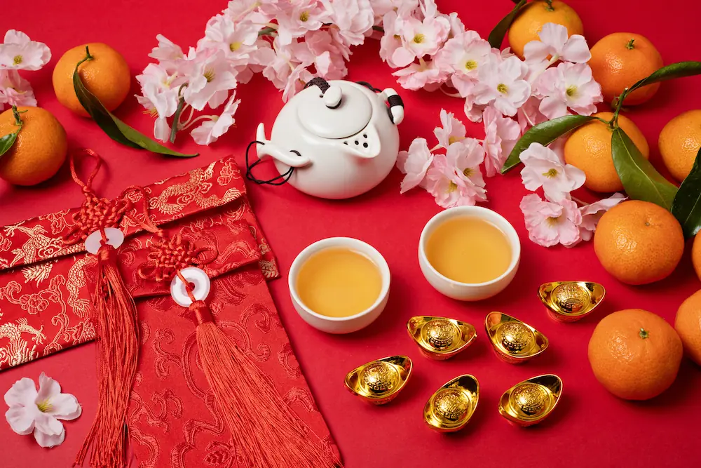 Chinese new year festival decoration over red background. Traditional lunar new year flat lay with green tea, red envelopes, gold ingots with text meaning fortune, prosperity, wealth. Top view