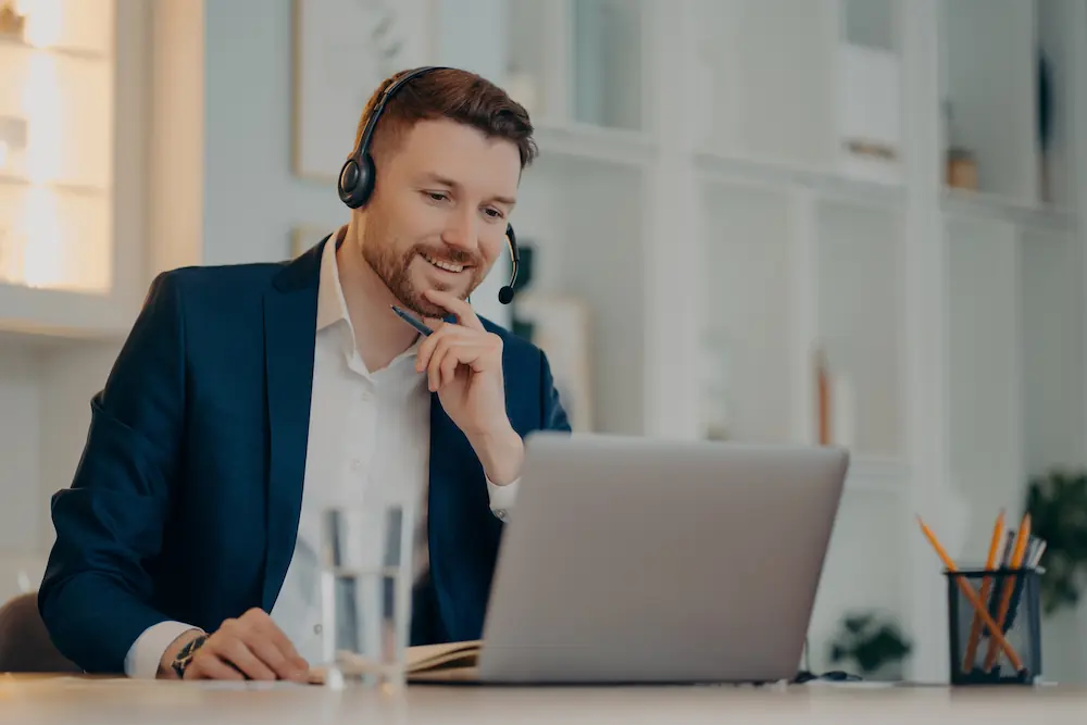 Professional male operator works in callcenter uses headset and laptop computer talks on video call or virtual webcam event wears formal clothes poses against cozy interior has online meeting